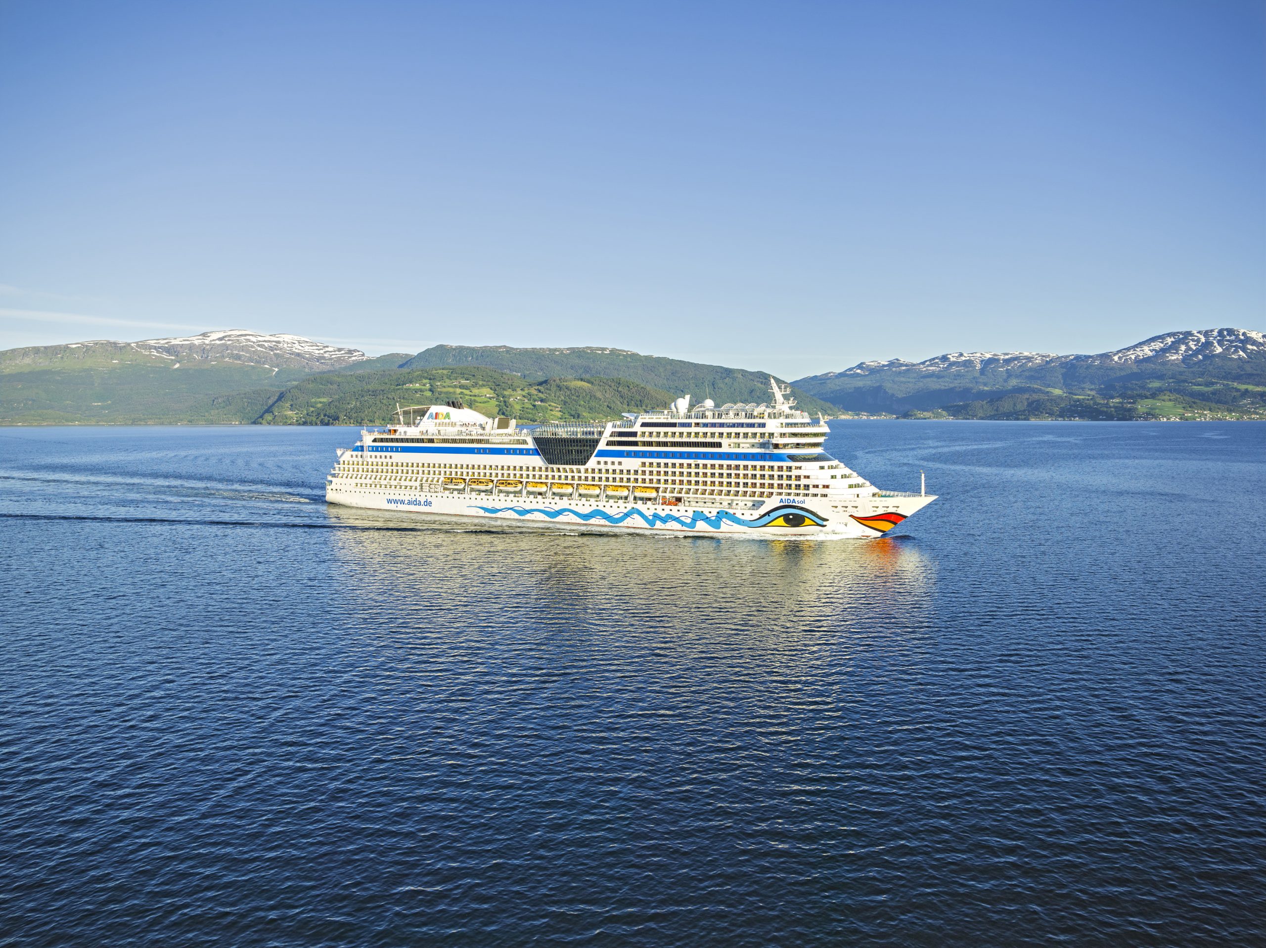 Das Kreuzfahrtschiff AIDAsol fährt bei klarem Wetter durch einen ruhigen Fjord. Im Hintergrund sind grüne, hügelige Landschaften mit schneebedeckten Berggipfeln zu sehen. Der Himmel ist blau und wolkenlos.