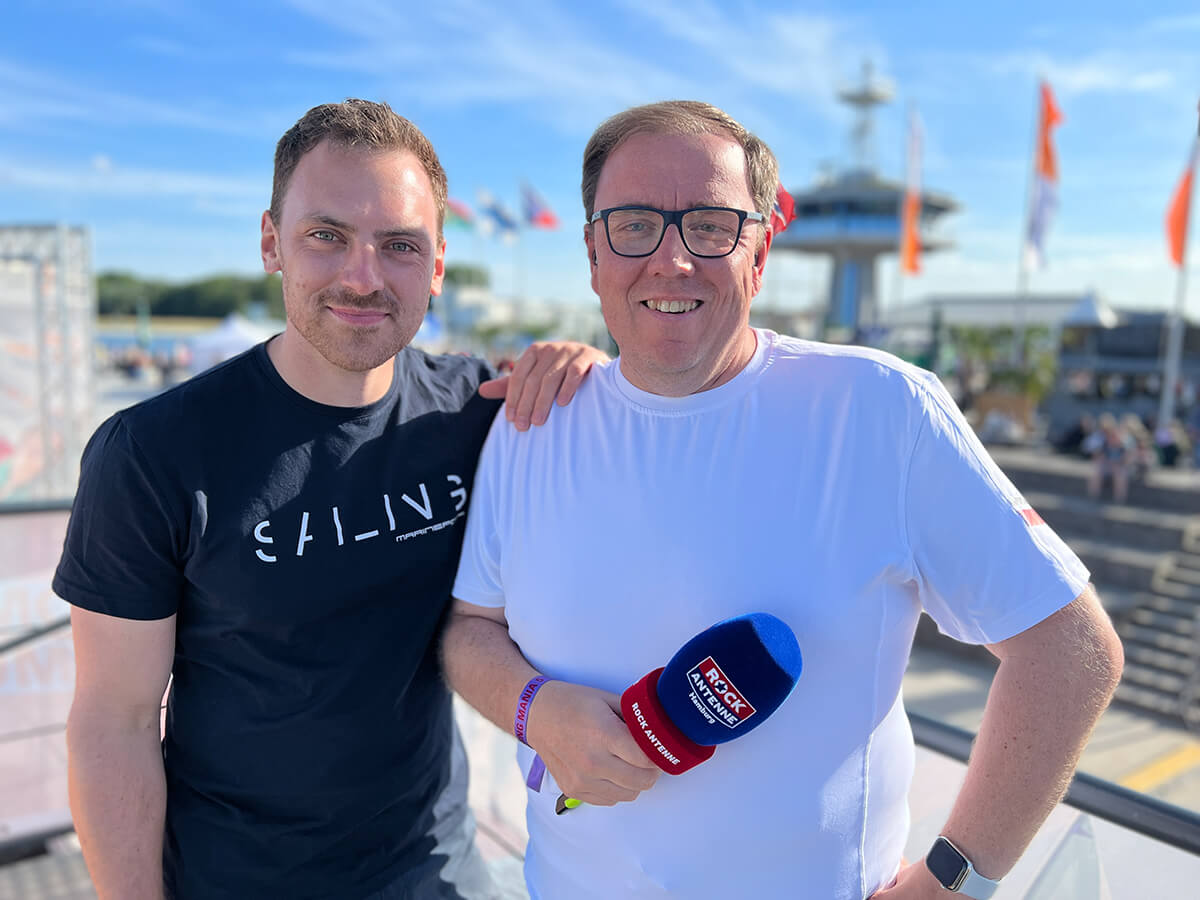 Jascha Bergmann und Sven Flohr, Moderatoren der Hamburg Cruise Days Live Sendung, stehen an einem Hafen mit Mikrofon und lässiger Kleidung