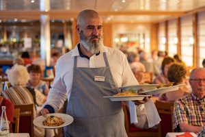 Kellner trägt graue Schürze und serviert zwei Teller mit Mittag- und Abendmenüs in einem belebten Restaurant