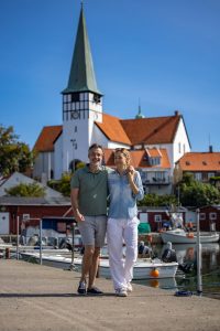 Paar spaziert am Hafen mit Booten und Kirche mit spitzem Turm im Hintergrund bei klarem Himmel