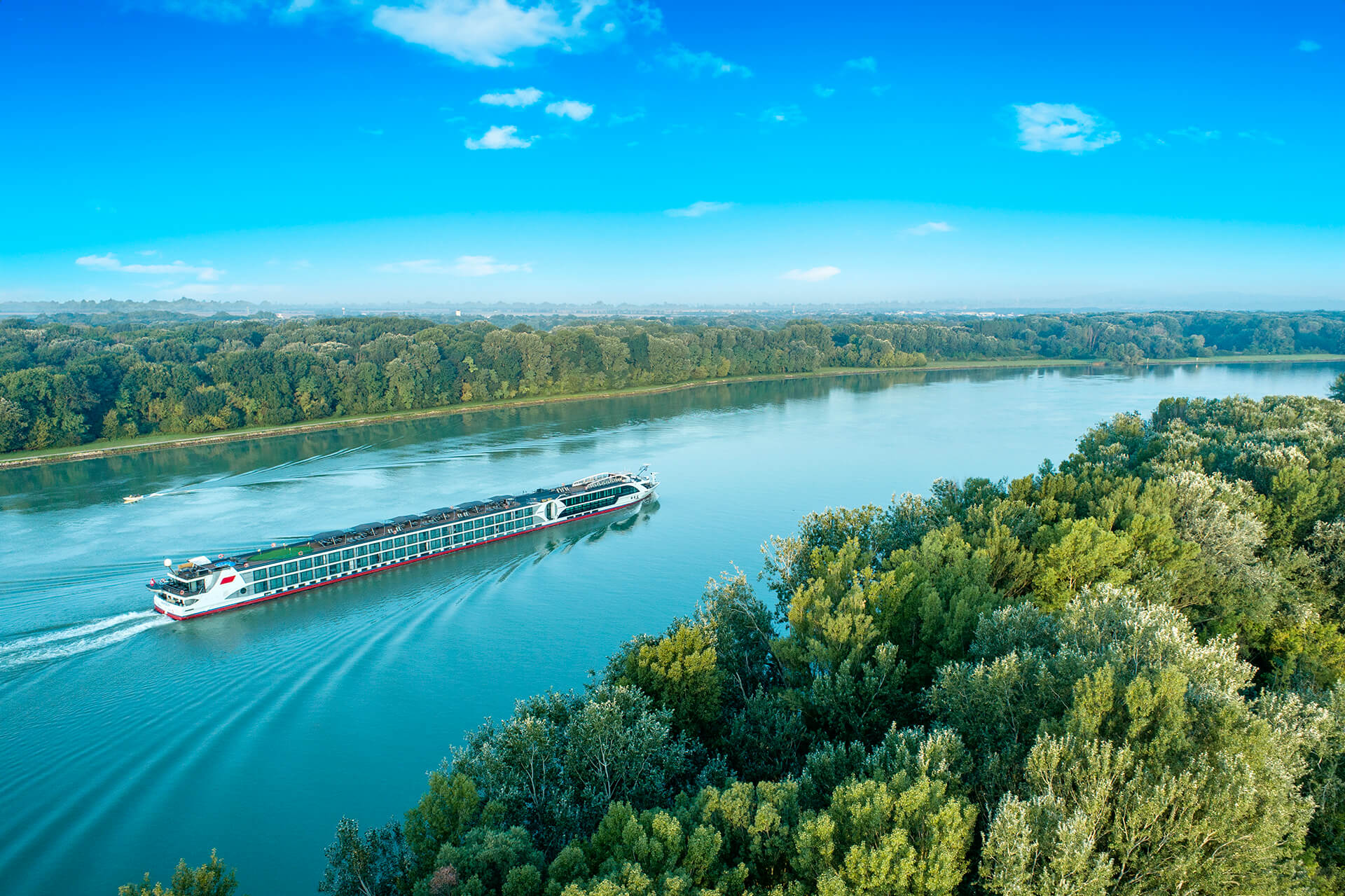 Flusskreuzfahrtschiff nickoVISION fährt auf der Donau, umgeben von grünen Uferwäldern unter blauem Himmel