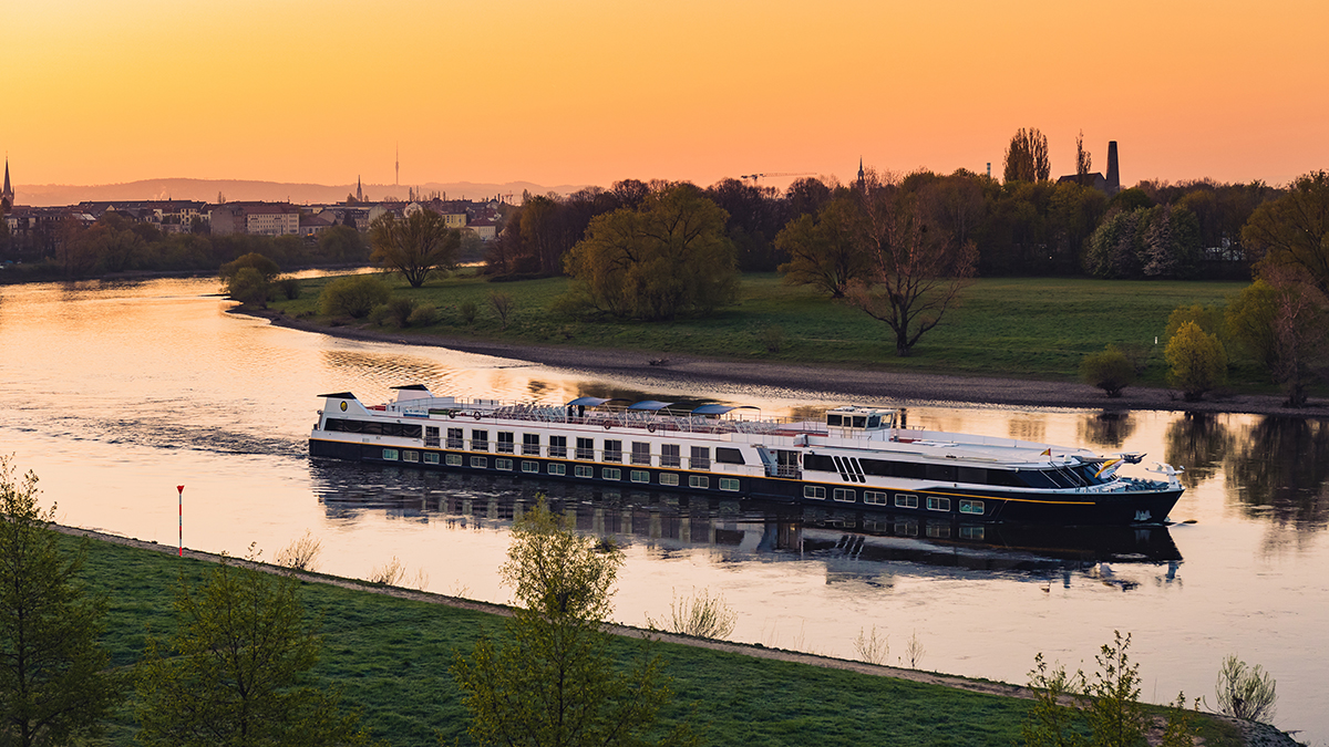 Das Flusskreuzfahrtschiff MS Sans Souci fährt bei Sonnenuntergang auf einem ruhigen Fluss, umgeben von Wiesen und Bäumen nahe einer Stadt.