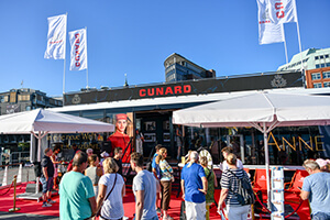 Menschen stehen vor einem Cunard-Stand mit weißen Zeltdächern und roten Sitzmöbeln bei sonnigem Wetter
