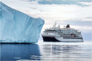 Kreuzfahrtschiff fährt auf ruhigem Wasser neben einem großen, blau-weißen Eisberg unter bewölktem Himmel