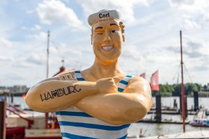 Eine Statue eines Matrosen mit gestreiftem Oberteil und tätowiertem Schriftzug 'HAMBURG' auf dem Arm. Im Hintergrund sind Boote und ein Hafen zu sehen.