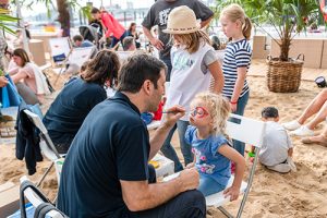 Ein Mann sitzt auf einem Stuhl und malt einem kleinen Mädchen im blauen Shirt das Gesicht. Im Hintergrund stehen mehrere Kinder und Erwachsene auf einem Sandboden.