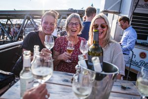Fünf Personen halten Gläser mit Getränken auf einer Terrasse. Im Hintergrund sind Menschen und ein Getränkewagen sichtbar.