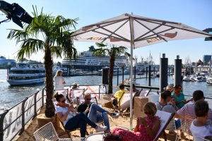 Eine Strandbar mit Palmen, Sonnenschirmen und Gästen, die auf Liegen sitzen. Im Hintergrund sind Kreuzfahrtschiffe und der Hafen von Hamburg sichtbar.
