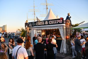 Ein Verkaufsstand mit dem Schriftzug 'FISH & CHIPS' und einem weißen Zelt. Menschen stehen in einer Schlange davor, im Hintergrund sind Segelschiffe und die Elbphilharmonie zu sehen.