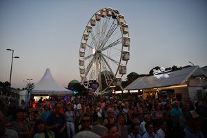 Eine große Menschenmenge steht vor einem Riesenrad bei Sonnenuntergang. Im Hintergrund sind ein Zelt und mehrere Gebäude sichtbar.