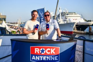 Zwei Personen stehen an einem Tisch mit 'ROCK ANTENNE'-Logo, halten Mikrofone. Im Hintergrund sind Kreuzfahrtschiffe und eine Menschenmenge sichtbar.