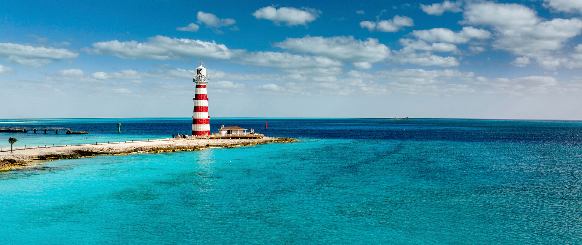 Leuchtturm mit roten und weißen Streifen am Ufer, umgeben von klarem blauem Meer unter bewölktem Himmel