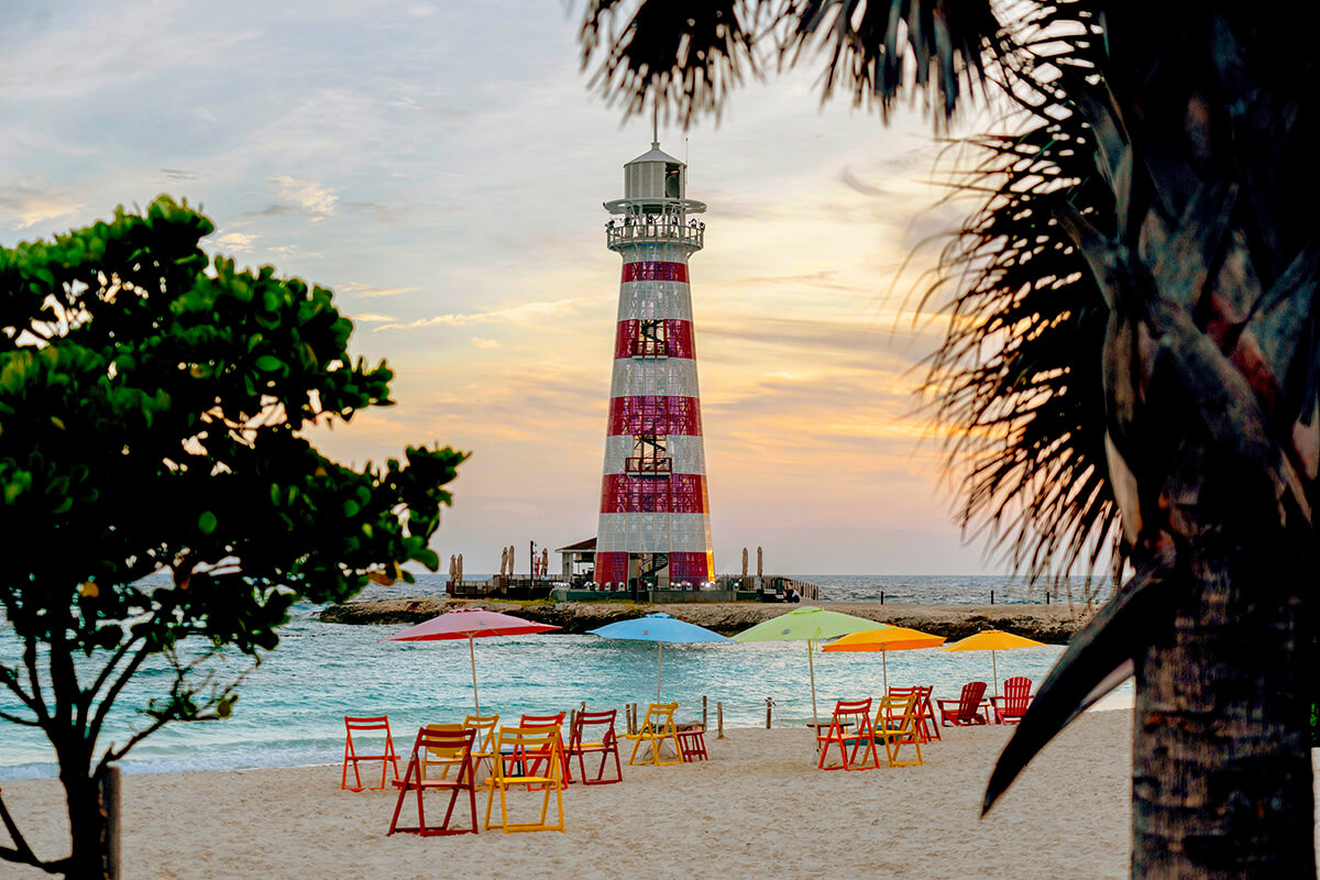 Leuchtturm mit roten und weißen Streifen am Strand bei Sonnenuntergang, bunte Sonnenschirme und Stühle im Sand
