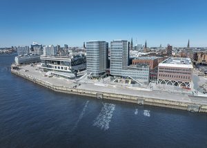 Luftaufnahme des Westfield Hamburg mit modernen Gebäuden am Wasser, Hafenanlagen und Stadtlandschaft im Hintergrund.