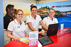 Drei Mitarbeitende in weißen Poloshirts stehen an einem roten Messetresen mit Laptop und Informationsmaterial vor einem Nicko Cruises Werbebanner