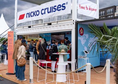 Messestand von Nicko Cruises mit Banner 'time to discover', mehreren Besuchern und Dekoration mit Palmen und Weltkarte