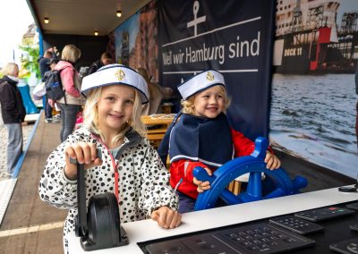 Zwei Kinder mit Matrosenmützen spielen an einem Steuerrad und Hebel in einem maritimen Ausstellungsbereich mit Banner 'Weil wir Hamburg sind' im Hintergrund.