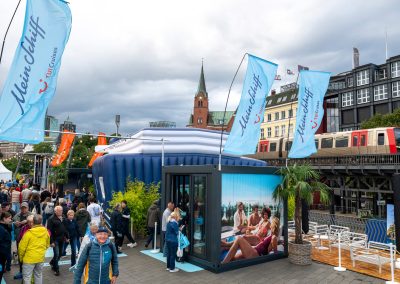 Menschenmenge vor blau-weißem, aufblasbarem Stand mit "Mein Schiff"-Fahnen und Bild von Personen am Strand, im Hintergrund S-Bahn und Kirchturm