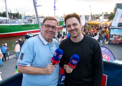 Zwei Männer stehen nebeneinander, halten blaue Mikrofone mit roten Aufsätzen und stehen an einem Hafen mit einem grünen Segelschiff und vielen Menschen im Hintergrund.