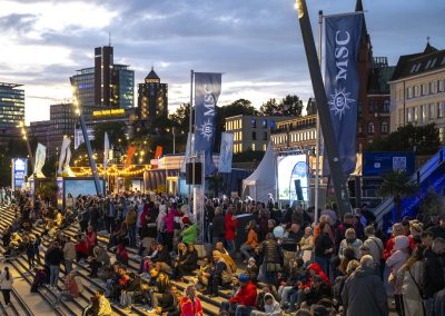 Menschenmenge auf Treppenstufen bei Abendveranstaltung am Hafen mit MSC-Fahnen und beleuchteten Gebäuden im Hintergrund.