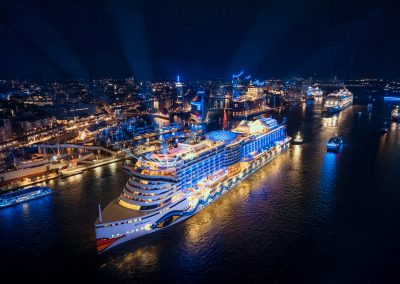 Beleuchtetes Kreuzfahrtschiff auf dunklem Wasser vor nächtlicher Hafenstadt mit weiteren Schiffen und Lichtern