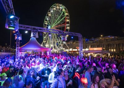 Große Menschenmenge bei Nachtveranstaltung vor beleuchtetem Riesenrad und Zelt mit Bühnenbeleuchtung