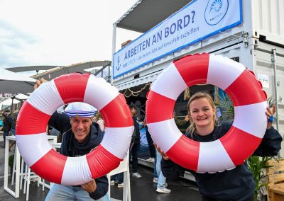Zwei Personen halten je einen rot-weiß gestreiften Rettungsring vor sich, im Hintergrund ein Container mit Banner 'ARBEITEN AN BORD?'.