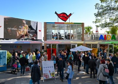Menschenmenge vor einem modernen, zweigeschossigen Messestand mit Glasfront, buntem 'Hi AIDA'-Schriftzug und rotem Lippen-Logo auf dem Dach