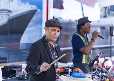 Zwei Musiker, einer spielt Schlagzeug mit Drumsticks, der andere singt in ein Mikrofon, im Hintergrund ein Schiff und Wasser.