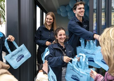 Mehrere Personen in dunkelblauen Jacken verteilen hellblaue Taschen mit dem Schriftzug 'Mein Schiff' und einem Schiff-Logo an Menschen im Vordergrund.