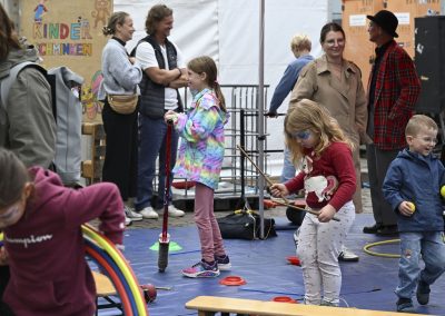 Kinder spielen mit verschiedenen Spielgeräten auf einer Matte, im Hintergrund steht ein Schild mit der Aufschrift 'Kinder schminken'.