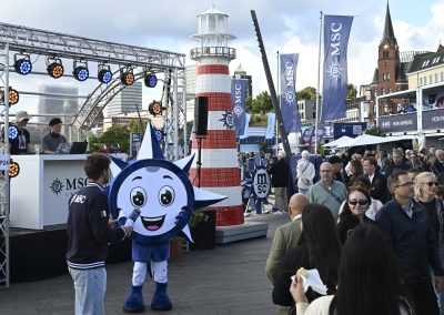 Menschenmenge auf einem Platz mit MSC-Fahnen, einem großen rot-weiß gestreiften Leuchtturmmodell und einer blauen Maskottchenfigur mit lachendem Gesicht.