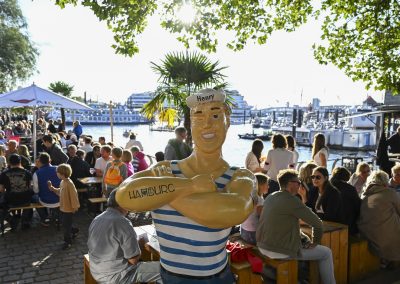 Figur eines Matrosen mit gestreiftem Hemd und Schriftzug 'Hamburg' auf dem Arm, im Hintergrund Menschen an Tischen am Wasser bei Sonnenschein.