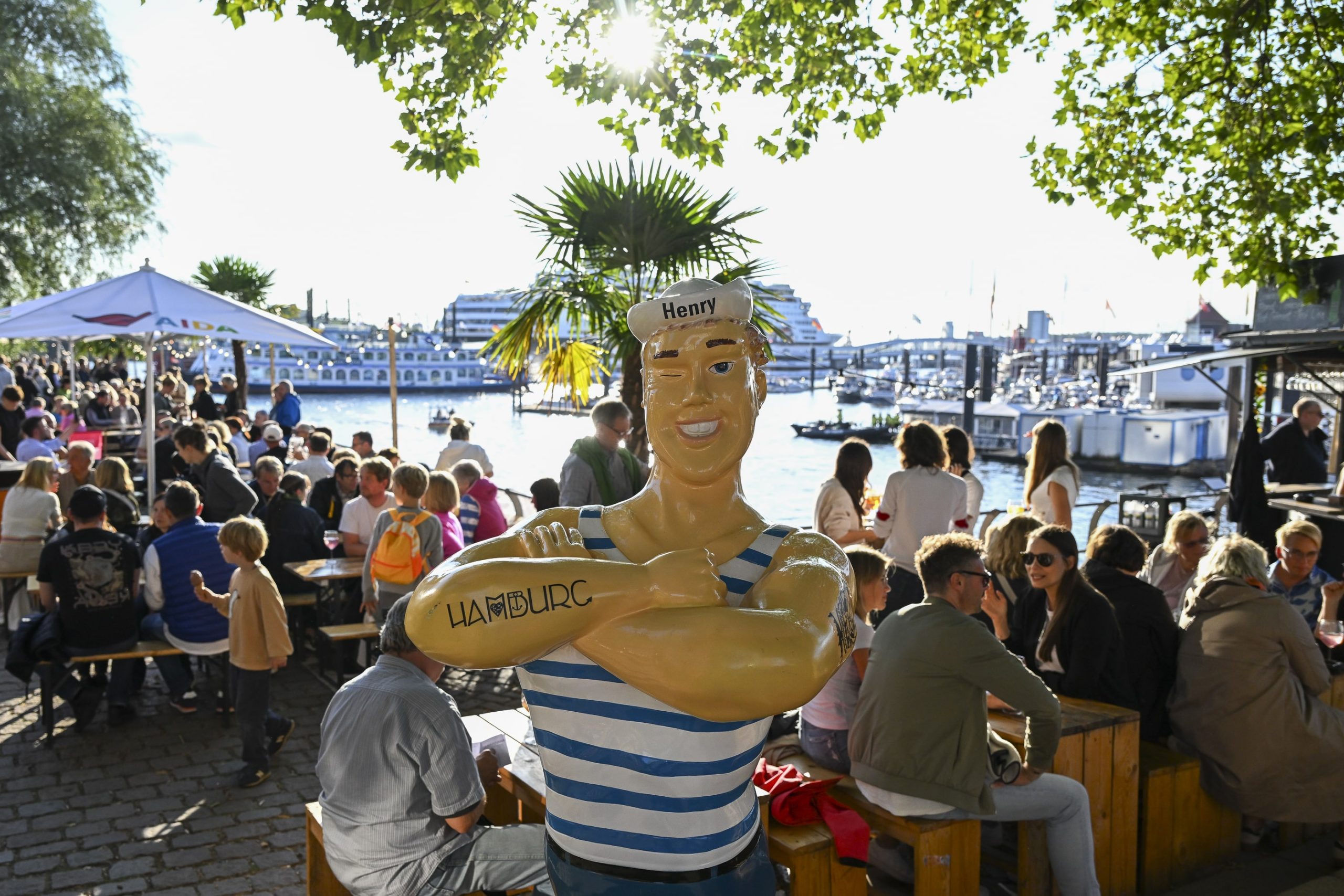 Figur eines Matrosen mit gestreiftem Hemd und Schriftzug 'Hamburg' auf dem Arm, im Hintergrund Menschen an Tischen am Wasser bei Sonnenschein.
