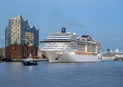 Kreuzfahrtschiff im Hafen vor der Elbphilharmonie in Hamburg bei Dämmerung, daneben mehrere kleinere Boote auf dem Wasser.