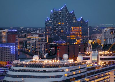 Kreuzfahrtschiff im Hafen vor der Elbphilharmonie in Hamburg bei Abenddämmerung mit beleuchteten Fenstern und Gebäuden.
