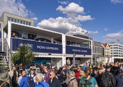 Menschenmenge vor zweistöckigem MSC Cruises-Stand mit Bannern 'MOIN HAMBURG' und 'MOIN MSC CRUISES' unter blauem Himmel mit Wolken.