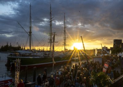 Segelschiff mit drei Masten im Hafen bei Sonnenuntergang, davor eine Menschenmenge am Ufer