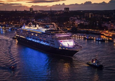 Kreuzfahrtschiff mit blau beleuchtetem Rumpf fährt bei Sonnenuntergang in einen Hafen, begleitet von mehreren kleineren Booten.